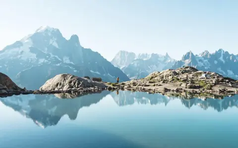 Un lac alpin reflète des montagnes majestueuses, un aperçu des plus belles randonnées de France. Une personne se tient sur la rive rocheuse.