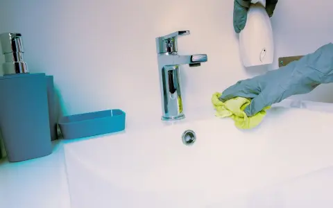 Manos con guantes limpian lavabo. Se usa un paño amarillo y un dispensador blanco.