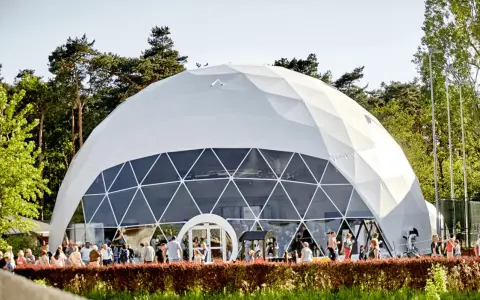 Una gran cúpula estructura blanca, con ventanas triangulares, se alza en un día soleado. Gente pasea frente a ella, rodeada de árboles verdes.
