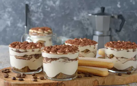 Plusieurs verrines de tiramisu sans café, garnies de cacao, côtoient des biscuits à la cuillère et des grains de café sur une planche en bois.