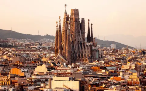Vue panoramique de Barcelone au coucher du soleil, avec la Sagrada Familia au centre. Les travaux de la Sagrada Familia continuent, sa silhouette imposante se détachant sur le ciel orangé.