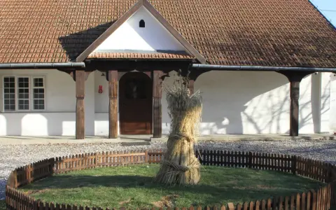Bronowicka chata symbol: snop zboża przed tradycyjnym domem z drewnianymi elementami i ceramicznym dachem.