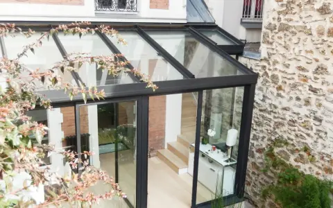 Une pergola transformée en véranda moderne, avec une structure en verre et métal, s'intégrant à une façade en pierre et brique.