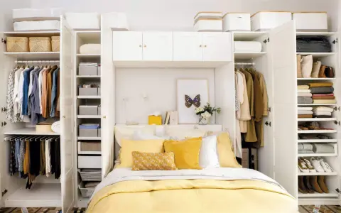 Habitación matrimonio pequeña con armario blanco, cama con ropa de cama amarilla y almohadas, y estanterías llenas de ropa y cestas.