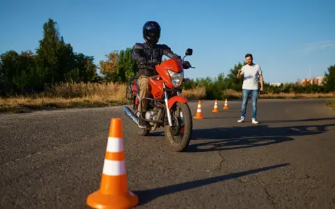 Persona con casco y equipo de moto naranja, practicando en circuito con conos. Un instructor observa. Preparándose para el carnet A.