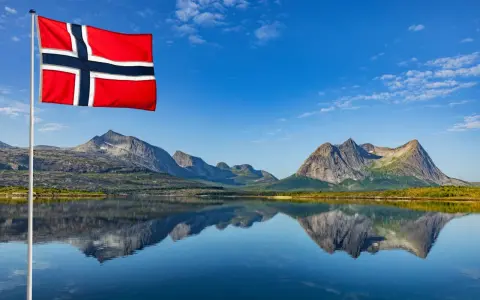 Norweska flaga powiewa nad jeziorem, odbijając się w wodzie. Czy do Norwegii potrzebny jest paszport? Piękne góry w tle.