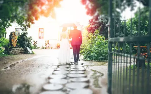 Para młoda idzie pod parasolem przez kamienną ścieżkę. W tle biały budynek i zielony ogród. To piękna brama weselna.