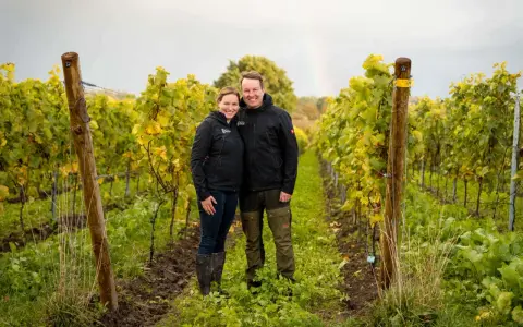 Ein Paar steht lächelnd in einem Weinberg in Schleswig-Holstein. Im Hintergrund ist ein Regenbogen zu sehen.