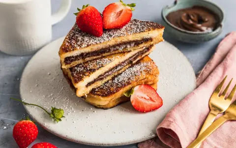 Gestapelte French Toast mit Nutella, bestäubt mit Puderzucker und garniert mit frischen Erdbeeren. Ein Glas Milch und eine Schüssel Nutella im Hintergrund.
