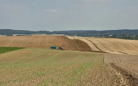 Czerwony traktor z przyczepą nawozi pole, przygotowując je do dalszej rekultywacji. W tle widać kolejne pola i las.