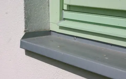 Szary parapet zewnętrzny zamontowany przy oknie z zielonymi okiennicami. Idealny moment, kiedy montować parapety zewnętrzne, to po ociepleniu elewacji.