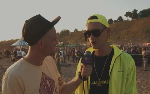 Dwóch mężczyzn na plaży, jeden z mikrofonem Red Bull, w tle tłum ludzi i scena.
