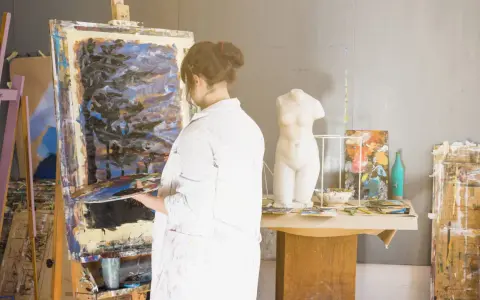 Un artisan artiste, en blouse blanche, tient une palette devant une toile abstraite dans son atelier. Une statue de femme est visible.