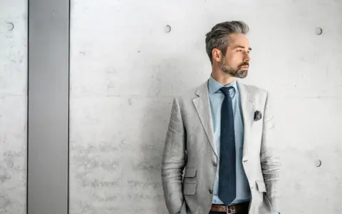 Stilvolles herren outfit sportlich elegant ab 50: Mann mit grauem Sakko, blauem Hemd und Krawatte vor Betonwand.