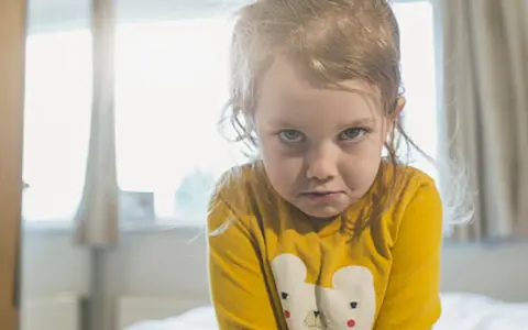 Mała dziewczynka w żółtej bluzce z misiem, z miną wyrażającą bunt 4 latka.