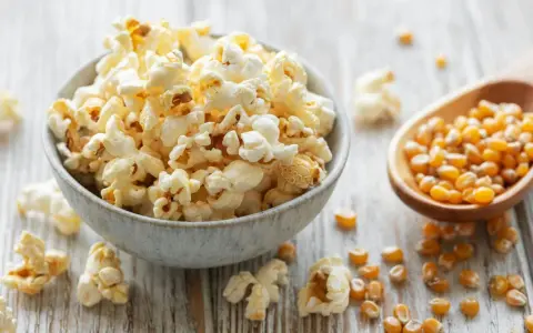 Schüssel mit frisch zubereitetem Popcorn im Topf, daneben eine Holzschaufel voller Maiskörner.