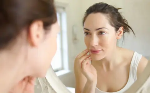 Une femme examine son visage dans un miroir, touchant sa joue. Sa peau semble très fine, peut-être signe d'une carence.