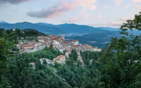 Borgo pittoresco su un colle, con case dai tetti rossi e un campanile, incastonato tra montagne e alberi. Via Francisca del Lucomagno, un incanto.