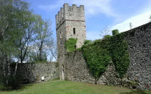 Antica torre del forte di Orino, circondata da mura in pietra e vegetazione rigogliosa, sotto un cielo azzurro.