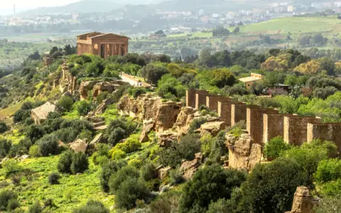 Ancient Greek temples stand proudly on a ridge in the Valley of the Temples, Sicily, surrounded by lush greenery and a distant town.