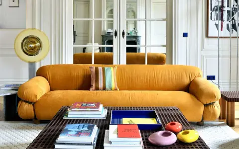 Divano senape con cuscino a righe colorate. Tavolino con libri e oggetti decorativi. Abbinamenti moderni e audaci.