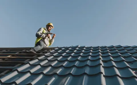 Murarz w kasku i kamizelce pracujący wkrętarką na dachu z niebieskiej dachówki.