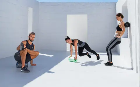 Drei Sportler nutzen eine **blackroll vor oder nach dem Training**. Ein Mann dehnt sich, eine Frau macht Mountain Climbers, die andere lehnt sich an die Wand.