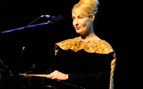 Kobieta w czarnej sukni z ozdobnym kołnierzem gra na instrumencie, tworząc muzykę. Kto śpiewa?