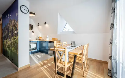 Przytulny apartament pod wierchem. W kuchni niebieskie szafki, jadalnia z drewnianym stołem. Na ścianie fototapeta z górami.