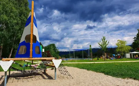 Spielplatz am Badesee Ludwigsburg mit Segel-Klettergerüst und Sandkasten. Im Hintergrund der See und Menschen.