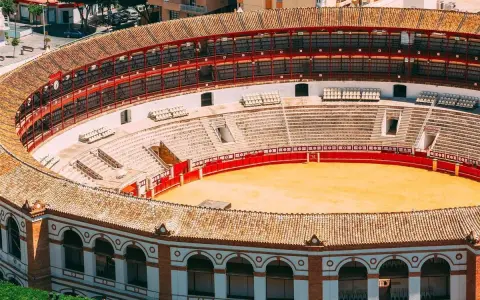 Leere Plaza de Toros mit roten Geländern und sandiger Arena. Die Sitzreihen sind leer, die Sonne scheint auf die traditionelle Architektur.