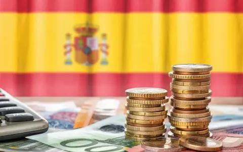 Monedas y billetes sobre una mesa, con la bandera de España de fondo. Una calculadora a la izquierda. Reflexión sobre qué son los tributos.