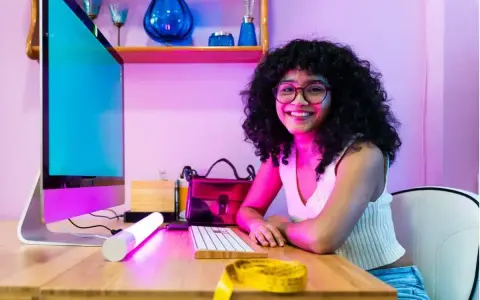 Giovane donna sorridente al computer, pronta a diventare e-commerce manager. Sul tavolo, un metro da sarta giallo.