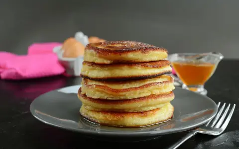 Pila di pancake senza latte, dorati e soffici, con sciroppo d'acero e uova sullo sfondo.