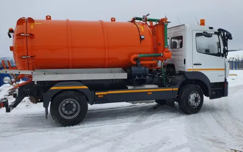 Pomarańczowy pojazd asenizacyjny z beczką i zielonymi wężami stoi na zaśnieżonej drodze.