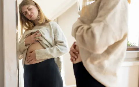 Ragazza si guarda allo specchio, toccandosi la pancia gonfia sopra i pantaloni stretti.