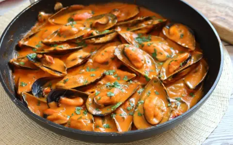 Mejillones con tomate en salsa cremosa, espolvoreados con perejil fresco, servidos en una sartén negra.