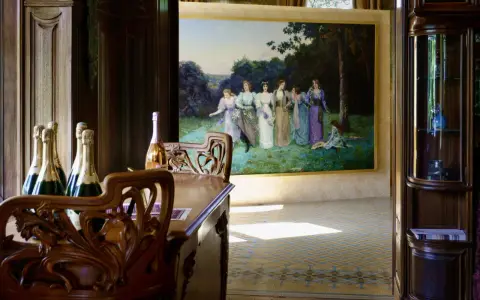 Bouteilles de champagne sur une table en bois sculpté, devant une peinture de femmes dans un jardin, ambiance Villa Mademoiselle Reims.