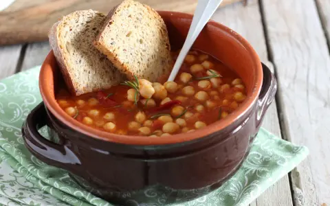 Una ciotola di terracotta con zuppa di ceci, pomodoro e rosmarino, accompagnata da fette di pane rustico.