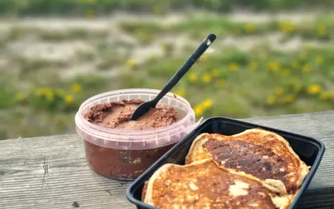 Färdiga proteinpannkakor i en svart behållare bredvid en burk med chokladpålägg, redo att avnjutas utomhus.