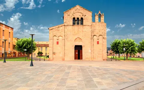 Piazza del centro di Olbia con la Basilica di San Simplicio, un'antica chiesa romanica in pietra chiara, sotto un cielo azzurro con nuvole bianche.