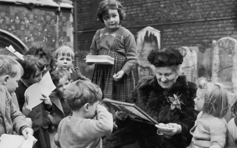 Maria Montessori joven, rodeada de niños, les muestra un libro. Una niña con tirantes sostiene una caja.