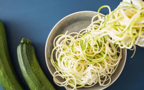 Espaguetis con calabacín recién hechos, listos para servir. Dos calabacines verdes reposan al lado de un plato lleno de fideos de calabacín.