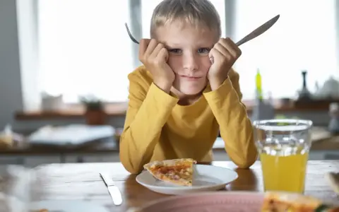 Chłopiec z widelcem i nożem w dłoniach, z miną mówiącą "dziecko nie chce jeść". Przed nim kawałek pizzy i szklanka soku.