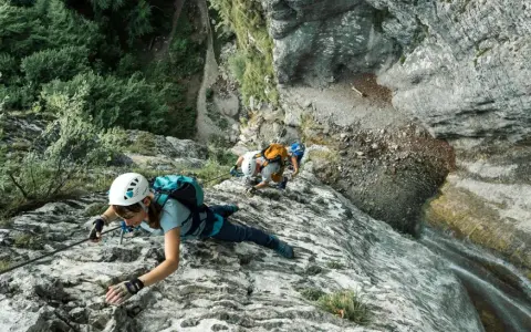 Osoby w kaskach i uprzężach pokonują skalną ścianę, używając liny i karabinków. Cały niezbędny via ferrata sprzęt zapewnia im bezpieczeństwo podczas wspinaczki.