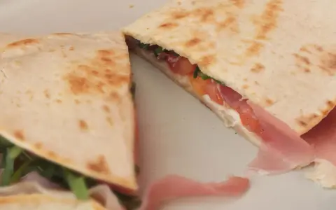 Pyszna piadina z szynką, pomidorami i rukolą, złożona na pół, prezentuje apetyczny przekrój.