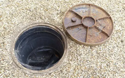 Inspection d'une canalisation bouchée sous la maison. Le couvercle rouillé est retiré, révélant le conduit noir.