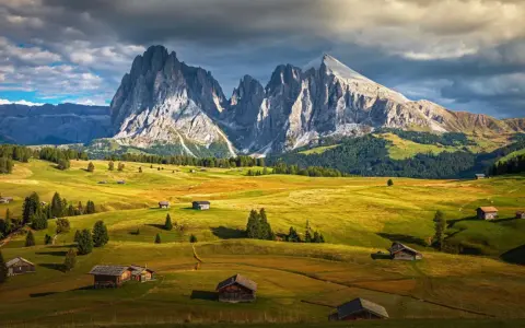 Un paesaggio alpino con un sasso lungo e imponente che domina un sasso piatto e verdeggiante, punteggiato da casette di legno.