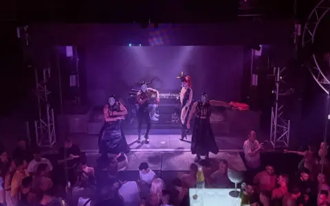 Performers in masks and elaborate outfits dance on stage at one of Valencia's vibrant gay bars, while a crowd watches below.