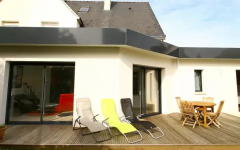 Extension de toit plat en bac acier, avec terrasse en bois, chaises longues et table de jardin.
